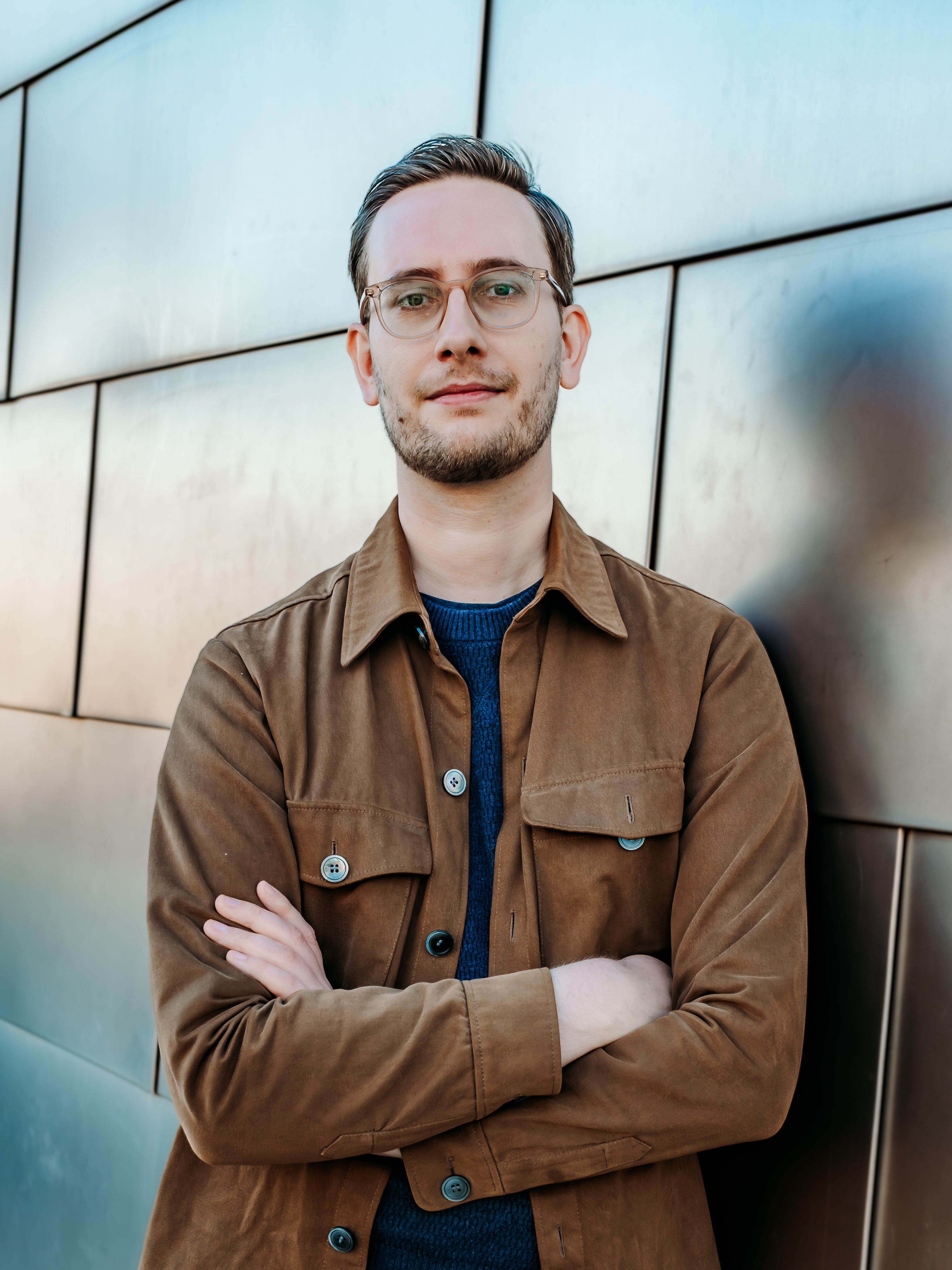 Portrait of Andrew Dana Hudson, facing the camera, in front of a metallic background. He has light skin, brown hair, and a short beard. He is wearing glasses, a blue sweater, and a brown shirt-jacket.